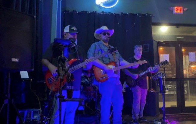 All four of the band on the stage at Pflutto's Tavern, the room's front doors and exit sign behind them.