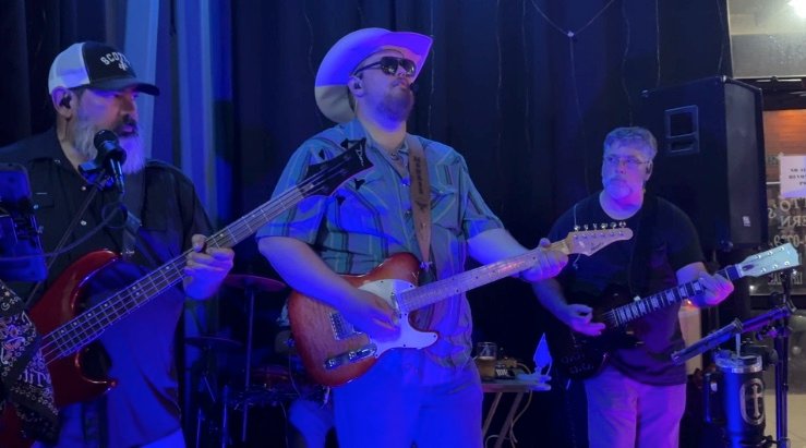 Lonesome Prayer mid-set at Pflutto's Tavern: the singer center stage in a white cowboy hat, bass player to his left and lead guitarist to his right under blue light.