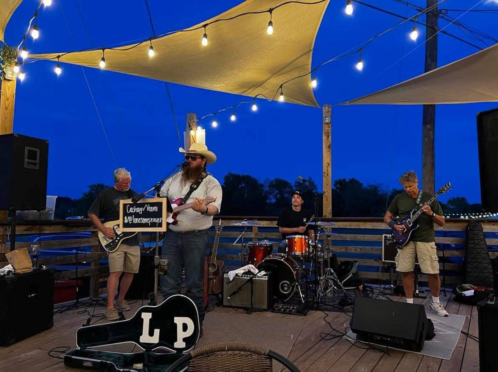 The band playing an outdoor deck stage at blue hour, string lights overhead and a guitar case painted LP set out front.