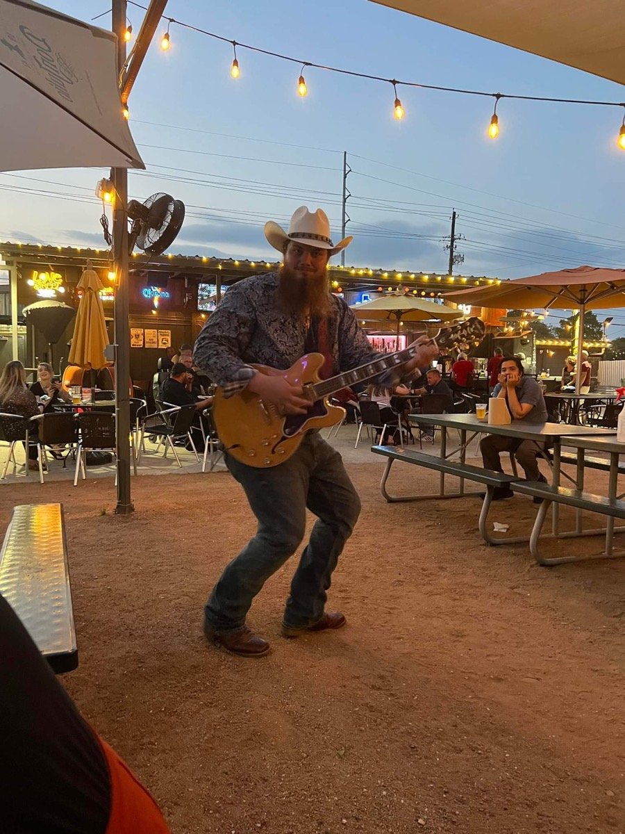 Guitarist playing on a gravel patio at blue hour with string lights and picnic tables.