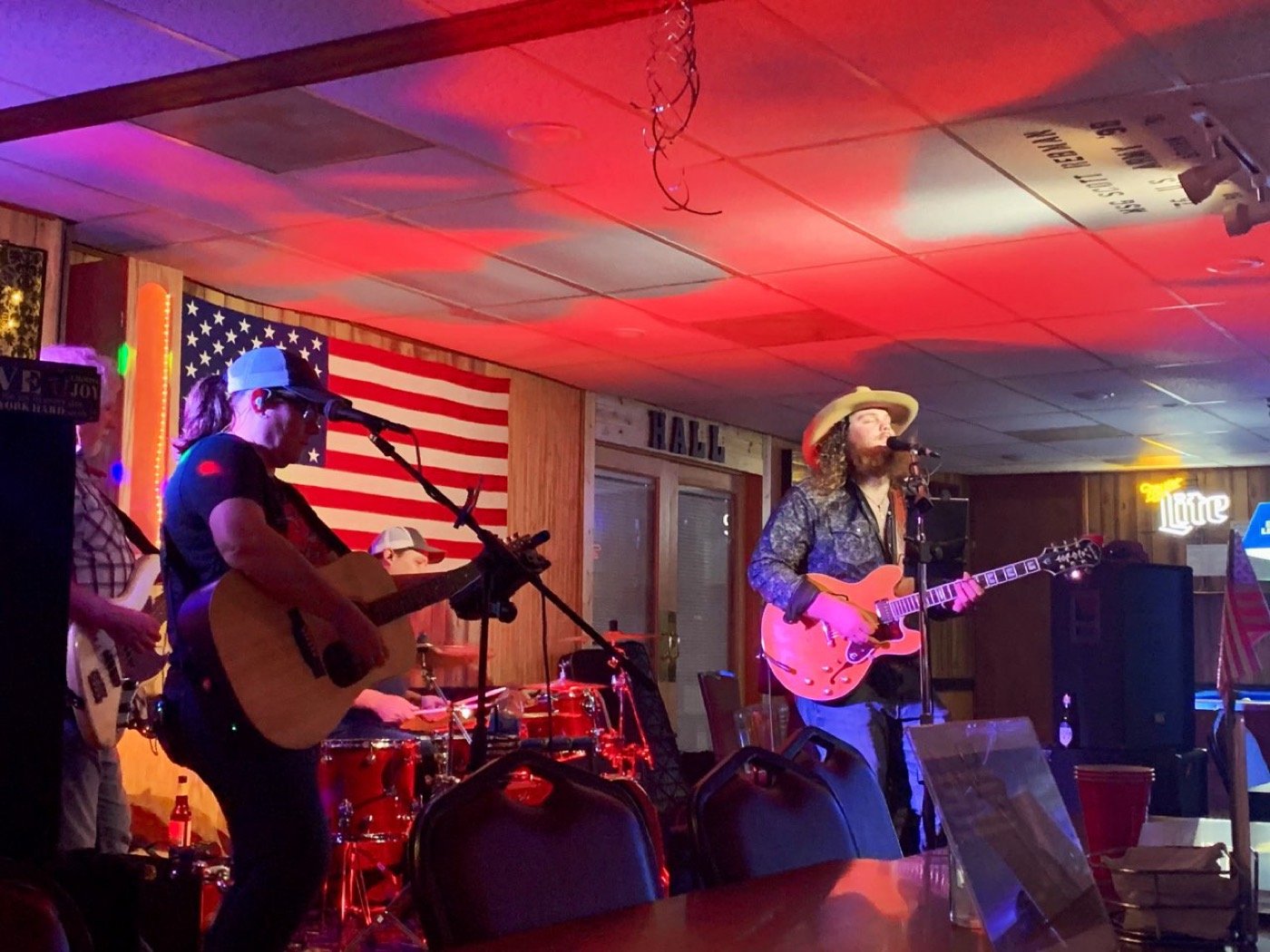 The band mid-set on an indoor stage washed in red light, flag on the wall behind them.